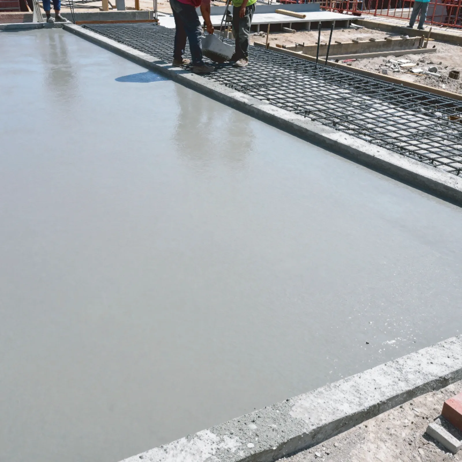 Two construction workers finishing the surface of a newly placed concrete slab, with reinforcing mesh laid out ready in the bay alongside