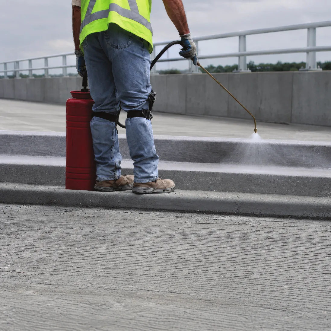 A worker in a hi-vis vest spraying liquid from a hand pump sprayer across a concrete deck, the fan of spray meeting the surface ahead of him