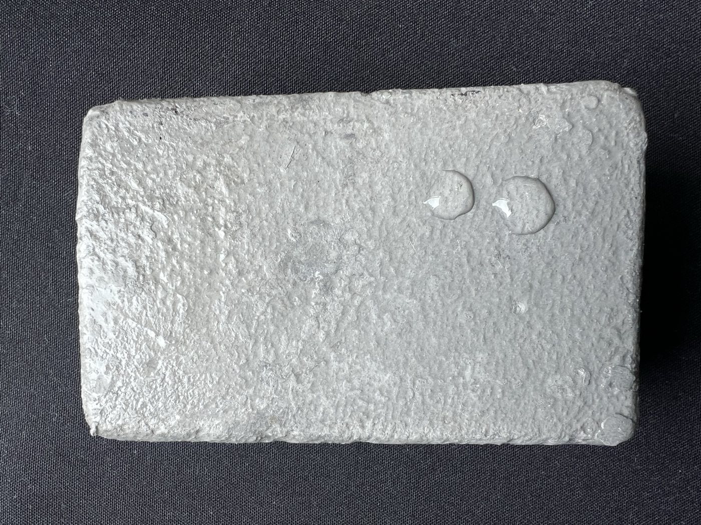A coated concrete sample on a dark cloth, its pale gray ceramic face textured across the whole surface, with two clear beads of water standing proud on it rather than soaking in
