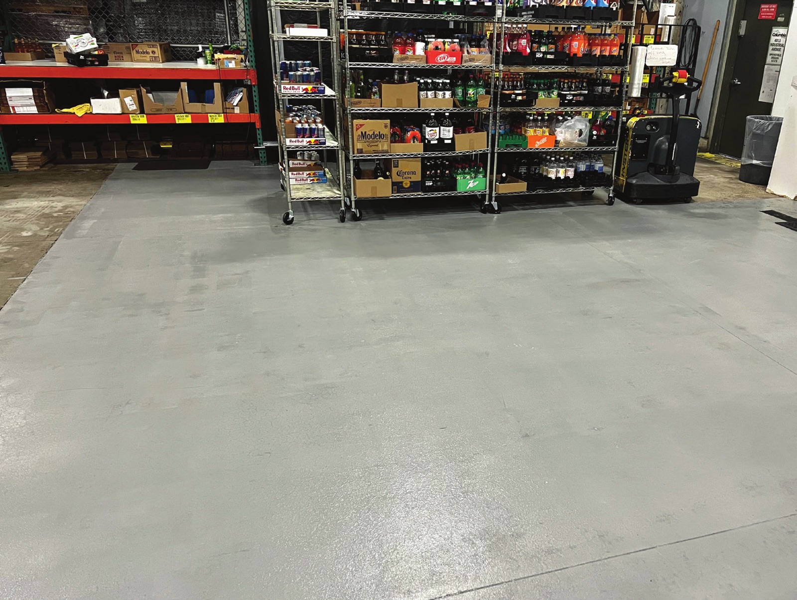 Rolling shelves stacked with bottled and canned drinks, pallet racking and an electric pallet truck standing on a gray coated concrete floor in a beverage distribution warehouse