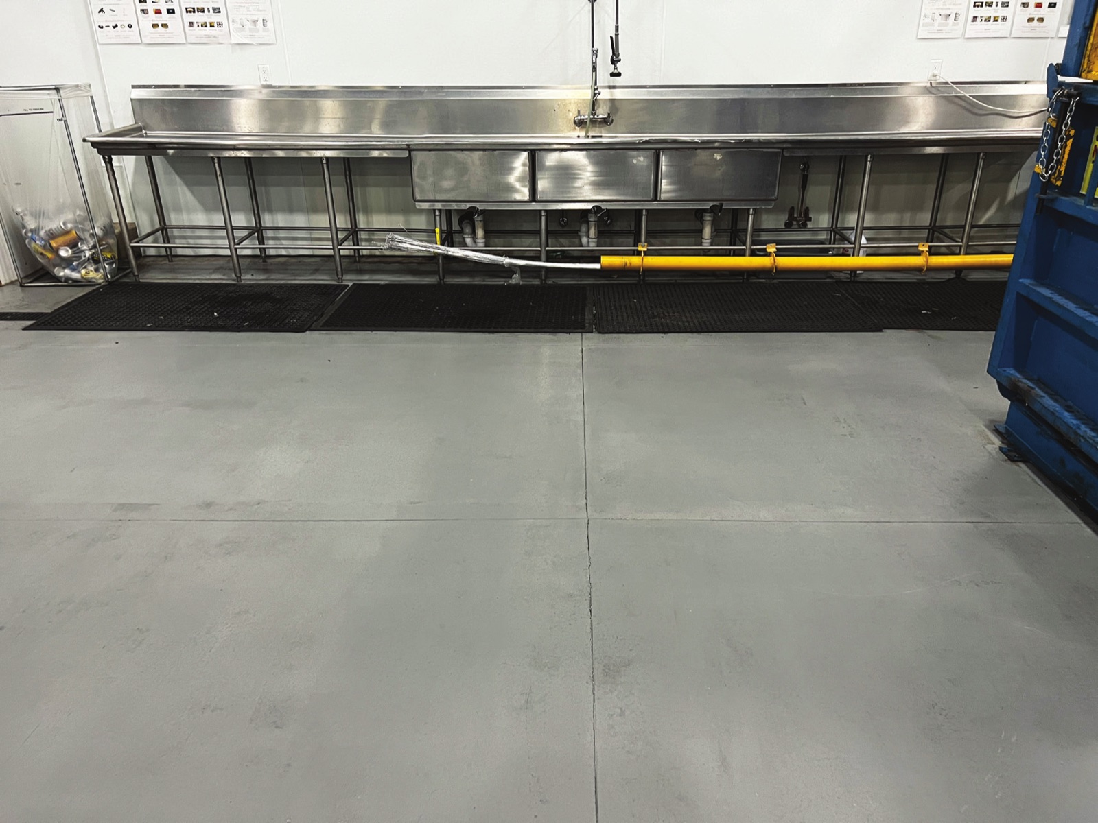 A long stainless steel wash-down sink along the wall of a food preparation room, with drainage matting and a gray coated concrete floor across the foreground