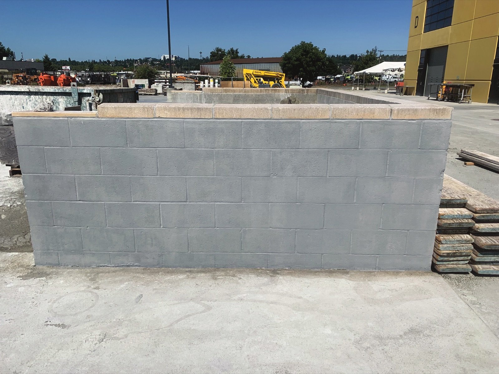 A concrete block wall in a contractor's yard finished in an even gray coating, with the untreated tan capping course left uncoated along the top
