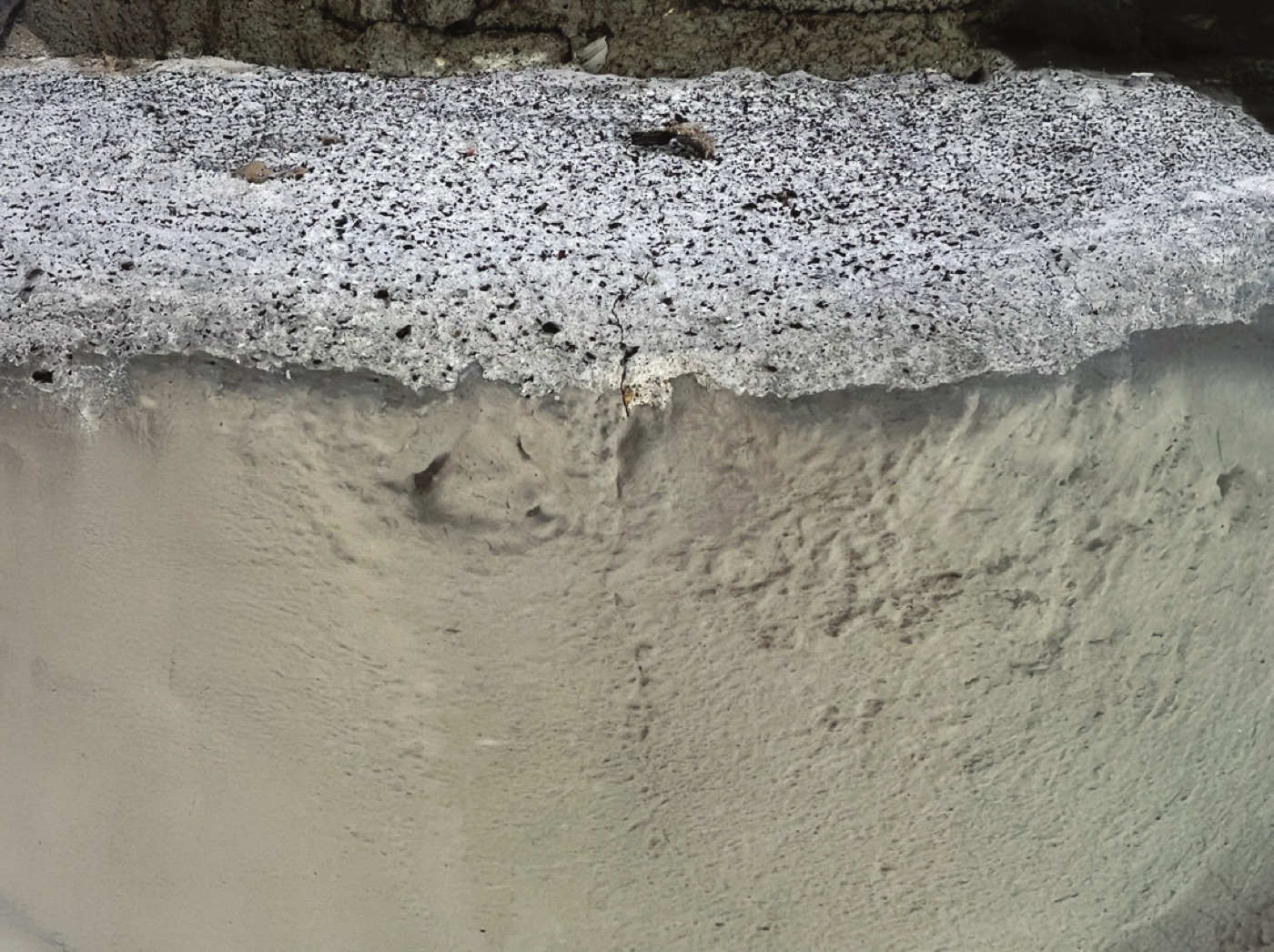 Close view of the edge of a concrete element, where a rough, open-pored top surface meets a smooth, dense pale face below it