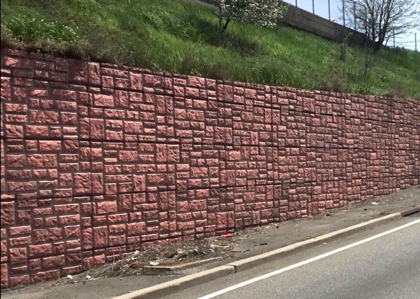 A long red patterned concrete highway retaining wall running beside a road shoulder, with a grassed bank rising behind it