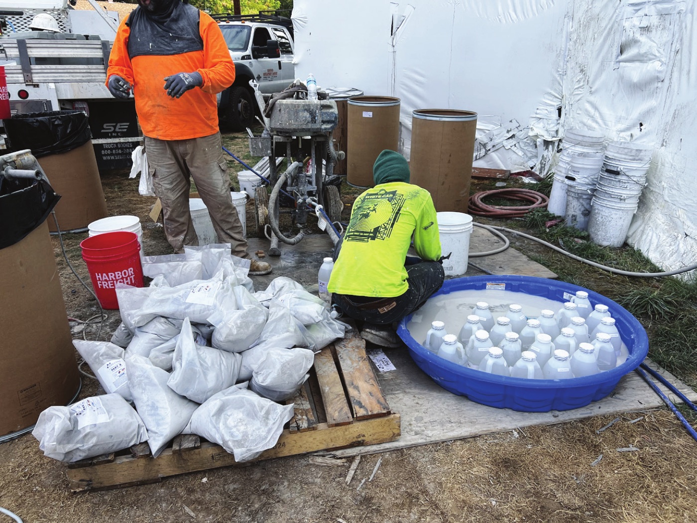 Two workers on a project site beside mixing equipment, with pallets of sealed powder sacks on one side and a tub of liquid containers on the other