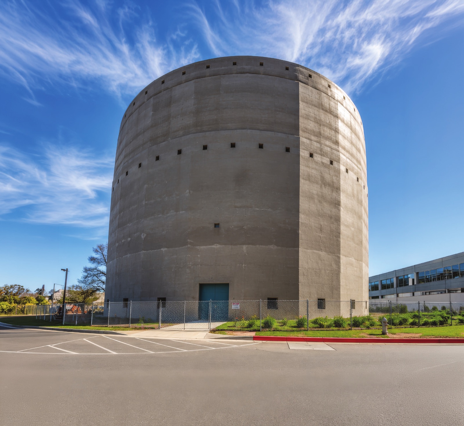 A large circular concrete water reservoir photographed against a blue sky, its treated wall surface even and unbroken from ground level to the roof line