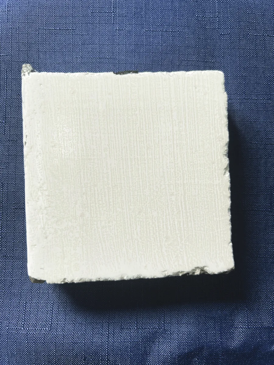 Square concrete sample with a white ceramic coating across its face, photographed on blue cloth