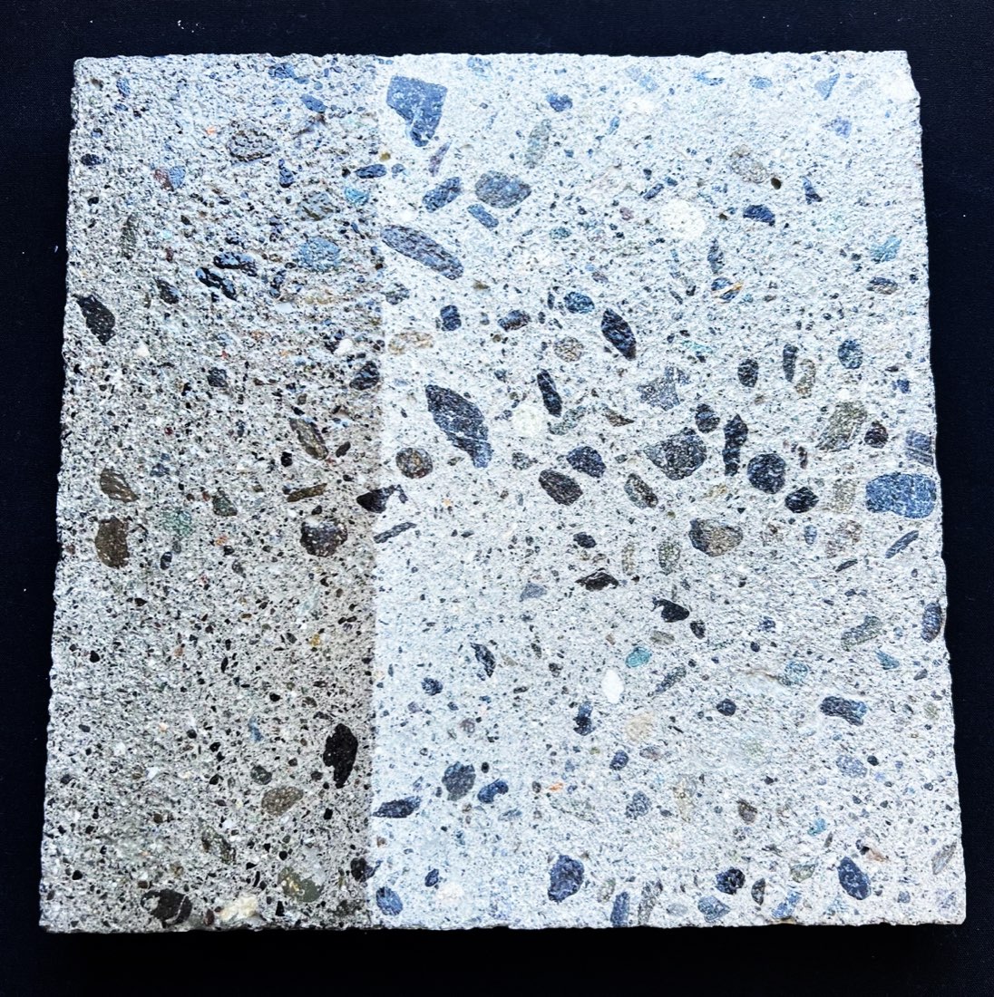 A square exposed-aggregate concrete slab on a dark cloth after wetting. A strip down the left side is visibly darker where water has soaked into the concrete, while the rest of the slab stays pale and dry