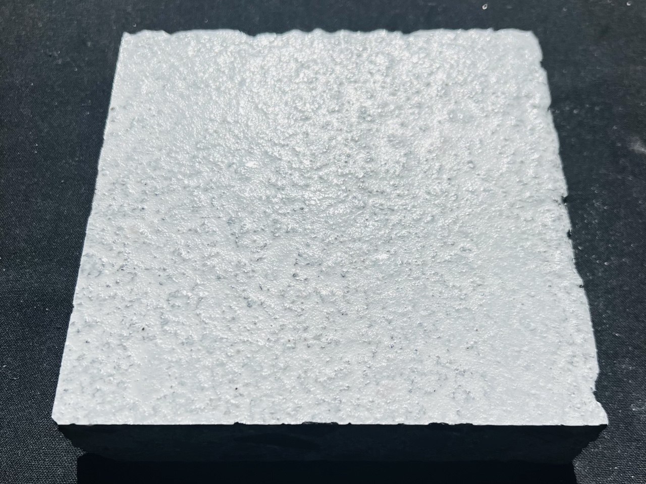 A square concrete sample on a dark cloth, its upper face coated in a bright white heavily textured ceramic finish with fine dark aggregate showing through, the uncoated dark edge of the block visible below