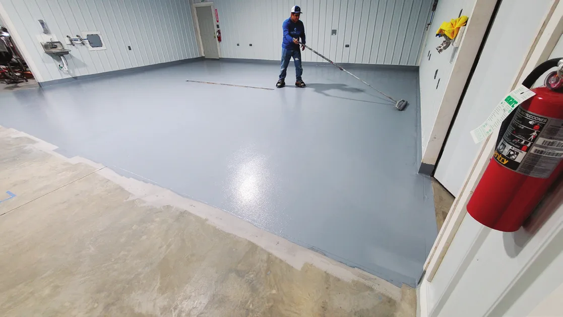 Applicator rolling a gray floor coating across a room with a long-handled roller, bare concrete still visible in the foreground