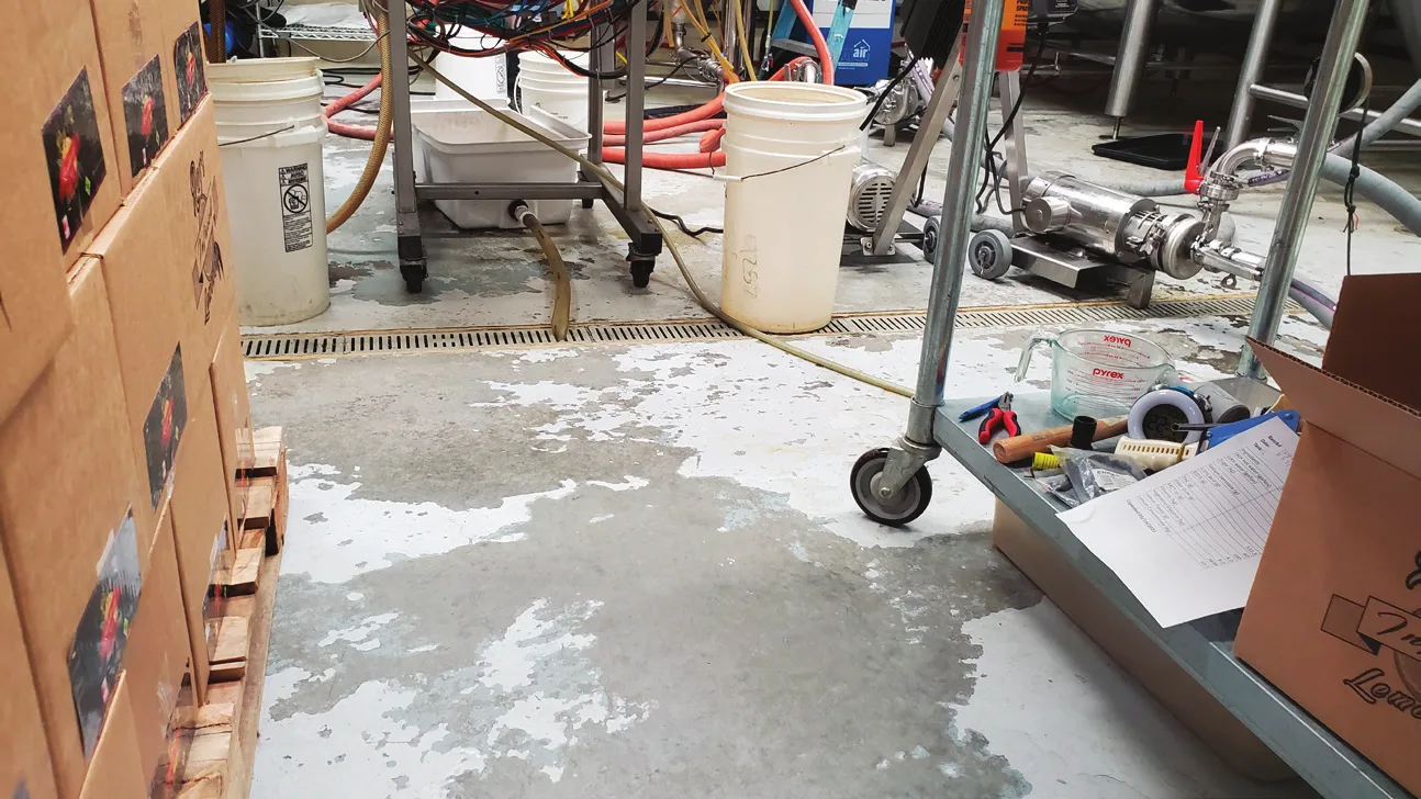 Failed floor coating peeling away in patches from bare concrete beside a trench drain in a beverage plant, with hoses, buckets and a stainless pump nearby