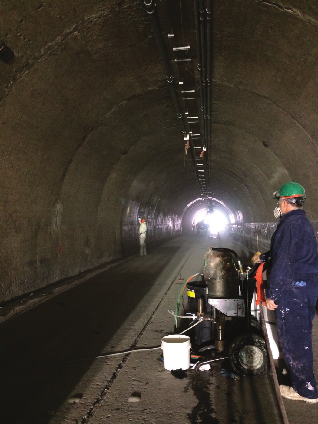 Two workers inside a concrete road tunnel, one in the distance spraying the arched tunnel lining and one in the foreground in a green hard hat and respirator operating a spray rig