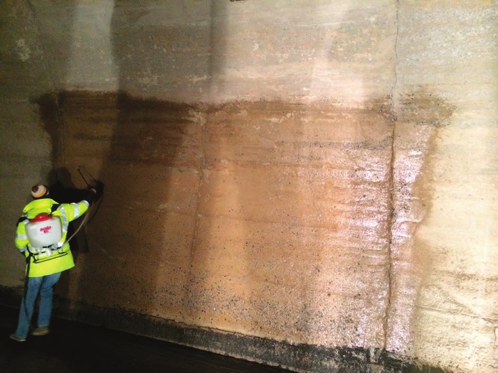 A worker in a high-visibility jacket with a backpack sprayer treating a large stained concrete wall beneath a structure