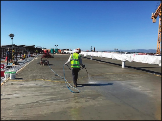 A worker in a high-visibility vest spraying liquid treatment across a large open concrete slab on a construction site under a clear sky