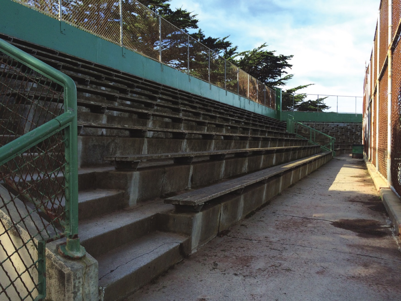 A weathered outdoor concrete grandstand with staining across the seating rows, green painted steel railings and a chain link fence